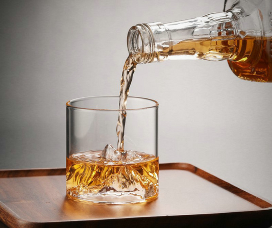 Whiskey being poured from a bottle into a mountain whiskey glass on a wooden tray with a grey background
