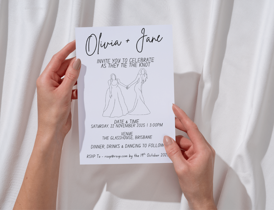 Hand holding a wedding invitation card with a lesbian couple illustration against a white curtain background.