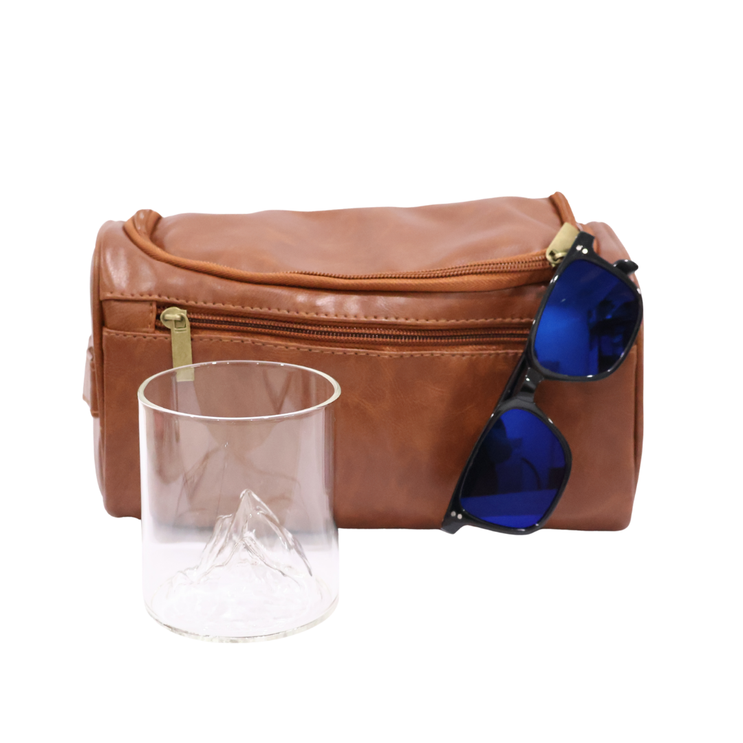 Groomsmen Gift Bundle consisting of a Brown leather toiletry bag with blue lens sunglasses and a clear mountain whiskey glass on a white background