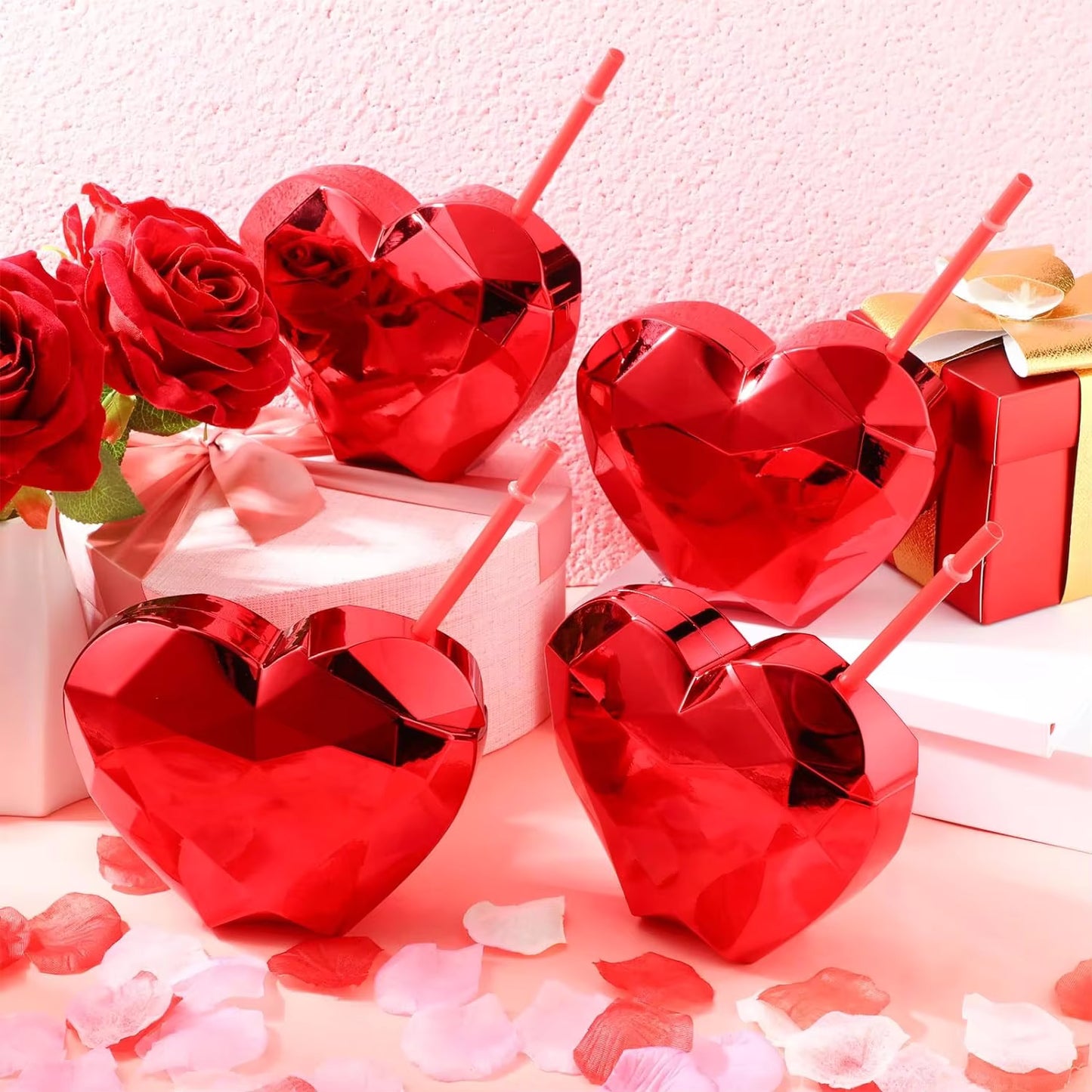 Red heart-shaped tumblers with straws on a romantic setting with roses and gift boxes.