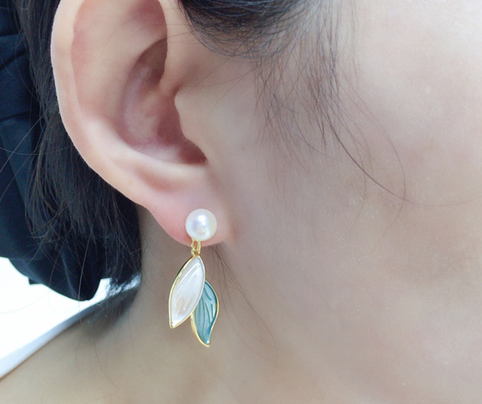 Close-up of an ear wearing a pearl earring with leaf-shaped gold and green charms.